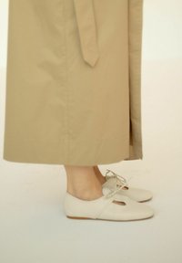 Scarpe flat beige con punta arrotondata, in materiale di pelle, con ritagli decorativi e dettaglio a fiocco, abbinate a una lunga gonna beige chiaro.
