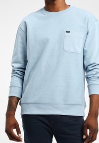 Ljusblå långärmad sweatshirt med bröstficka och liten svart "Lee"-etikett, bärs med mörka byxor, visar midja och armar.