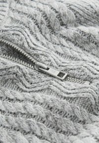 Gestrickter grauer Pullover mit hohem Kragen und Zickzack-Kabelmuster, mit einem silbernen Metallreißverschlussdetails am Kragen. Strukturiertes Material.