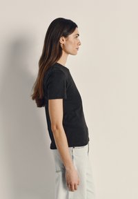 Jauna moteris su ilgais rudo plaukais, vilkinti juodą marškinėlį ir šviesiai mėlynas džinsines kelnes, stovinti šonu priešais neutralią šviesią foną.