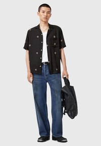 Ung mann med svart blomstermønstret kortermet skjorte, hvit tanktop, blå jeans, sorte støvler, og holder en svart lærryggsekk.