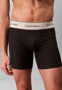 Manlig överkropp iförd svarta Calvin Klein kalsonger med beige midjeband och logotyp, stående mot en enkel grå bakgrund.