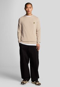 Pulover bej cu guler rotund, cu un logo galben, suprapus peste o cămașă albă, asortat cu pantaloni negri largi și adidași cu un model.