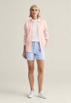 Kvinde iført hvide sneakers, blå blomstrede shorts, hvid strikket top og lyserød skjorte med knapper mod en neutral baggrund.