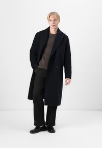 Laivastonsininen villakangastakki, jossa on kaksirivinen malli, viistot läpät ja teksturoitu kangas, päällä ruskea neulepaita ja mustat housut.