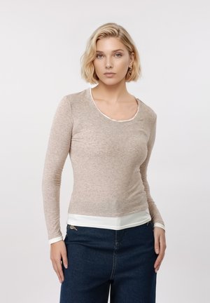 Ξανθιά γυναίκα με μπεζ πουλόβερ μακρυμάνικο πάνω από λευκό πουκάμισο και σκούρο μπλε τζιν, στέκεται μπροστά σε απλό ανοιχτόχρωμο φόντο.