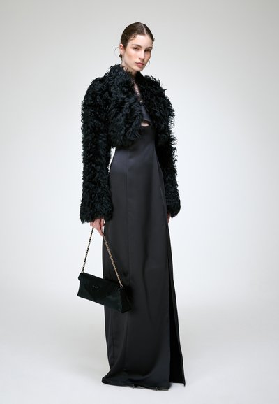 Femme portant une longue robe en satin noire avec une fente latérale, une veste courte noire en fourrure, et tenant un sac à main noir avec une anse chaîne dorée.
