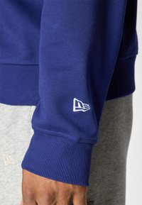 Marineblå sweatshirt med ribbede ærmer og et lille hvidt logo på venstre ærme, præsenteret mod lysegrå bukser.