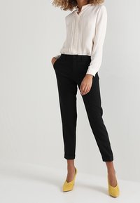 Bílá blůza s knoflíkovým zapínáním, černé tailored kalhoty a žluté lodičky na vysokém podpatku; hladká tkanina; kalhoty s manžetami; model pózuje s rukou na boku.