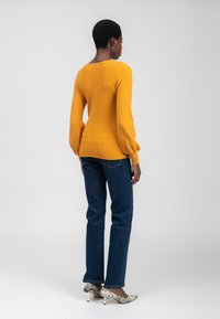 Frau, die mit dem Rücken zur Kamera steht und ein senfgelbes Langarmoberteil, dunkelblaue Jeans und weiße High Heels mit Schlangenledermuster trägt, vor einem einfarbigen Hintergrund.