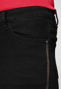 Mustat farkut ovat muotoilultaan kapeat, niissä on pieni etutasku ja koristeellinen vetoketjuyksityiskohta sivussa, ja ne ovat sileäpintaiset.