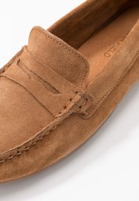 Mocassin en suède marron avec une couture, un léger pli au col et une semelle en caoutchouc plate, présenté sous un angle.
