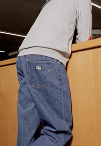 Fitted denim jeans in a medium wash with a straight leg design, featuring a back pocket logo patch and subtle stitching details.