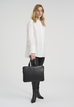 Femeie în picioare, purtând o cămașă albă cu mâneci lungi și pantaloni negri, ținând o servietă neagră din piele cu mânere, pe un fundal simplu.