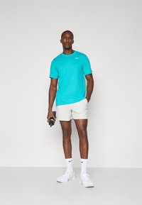 T-shirt Nike turchese, maniche corte, abbinata a pantaloni corti grigio chiaro. Il modello tiene in mano una borraccia nera, indossando sneaker e calze bianche.