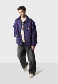 Fleece bunda v modro-červenom abstraktnom vzore, s gombíkovým zapínaním vpredu, náprsnej vreckom s logom, nosená cez svetlo béžový svetrový vzor, v kombinácii s tmavými džínsami.