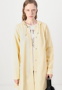 Chubasquero amarillo con capucha, que presenta cierres a presión y bolsillos laterales, usado sobre una blusa de color claro. Material suave y ligero.