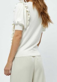 Vit stickad topp med volangshorts ärmar och ribbade fållar. Baksidan är slät och figurnära, kombinerad med ljusa byxor.