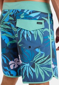 Herrarnas badshorts i blått med ett tropiskt mönster av gröna blad. Har en mintgrön midja och en bakficka med lock.