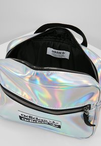 Bolso Adidas de color plateado iridiscente con un bolsillo frontal con cremallera, forro interior negro y etiqueta con el logo. Cuenta con un asa superior y detalles de cremallera en negro.