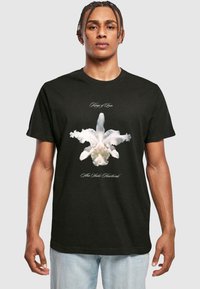 Sort bomulds-T-shirt med en hvid orkidé-grafik på fronten, med teksten "Kings of Leon" og "Alto Wake Heartbreak" i elegante skrifttyper.