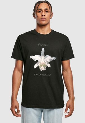 Sort bomulds-T-shirt med en hvid orkidé-grafik på fronten, med teksten "Kings of Leon" og "Alto Wake Heartbreak" i elegante skrifttyper.