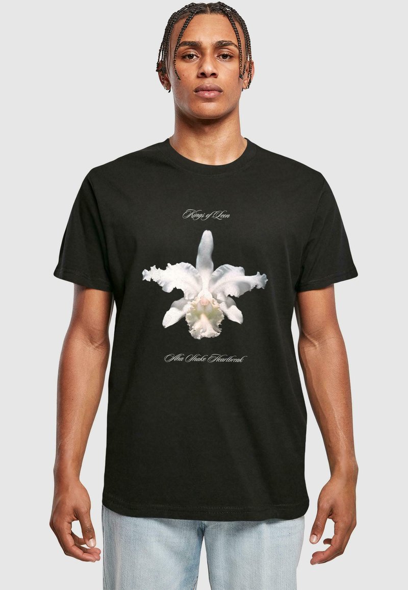Sort bomulds-T-shirt med en hvid orkidé-grafik på fronten, med teksten "Kings of Leon" og "Alto Wake Heartbreak" i elegante skrifttyper.