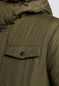 Olivgrön pufferjacka med hög krage, med dragkedjestängning, justerbar huva och en knappad bröstficka med svarta detaljer.