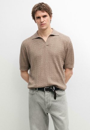 Mann trägt beige kurzärmeligen, kabelgestrickten Poloshirtpullover, in hellgraue Jeans mit einem schwarzen geflochtenen Gürtel gesteckt, und steht vor einer schlichten weißen Wand.