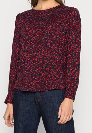 Kobieta w granatowej bluzce z długim rękawem w czerwony kwiatowy wzór, w połączeniu z ciemnoniebieskimi dżinsami.