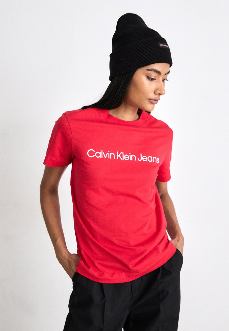 Červené bavlnené tričko s krátkymi rukávmi a okrúhlym výstrihom, s bielym textom "Calvin Klein Jeans". Nosí sa s čiernou pletenou čiapkou.