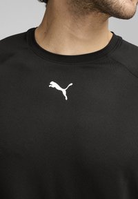 Juodas sportinis marškinėliai su lygiu paviršiumi. Priekyje yra baltas Puma logotipas. Apvalus iškirpimas ir trumpomis rankovėmis.