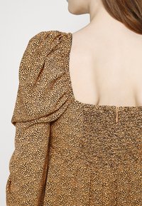 Robe imprimé léopard marron avec manches bouffantes, corsage smocké et dos décolleté. Comprend une fermeture éclair dissimulée et une jupe fluide.