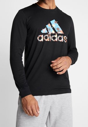 Homme portant un t-shirt Adidas noir à manches longues avec un logo thème sportif en couleurs pastel et un short gris clair sur fond uni.