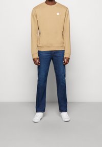 Felpa beige con maniche lunghe e collo a giro, abbinata a jeans blu scuro e sneakers bianche, in piedi contro uno sfondo semplice.