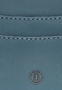 Getextureerde blauwe stof met fijne patronen, voorzien van een gestikte naad en een cirkelvormig logo met een boomontwerp dat in het midden onderaan is geperst.