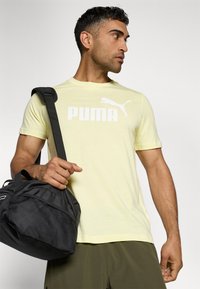 Light yellow short-sleeve T-shirt with white "PUMA" logo, paired with a black textured duffel bag and green shorts.