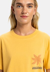 Gul bomulls T-skjorte med rund hals, korte ermer, og et trykt logo med blomsterdesign på venstre bryst.