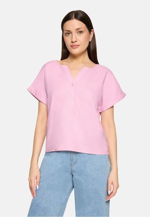 Kobieta o prostych ciemnych włosach nosi jasnoróżową bluzkę z krótkim rękawem na guziki oraz jasnoniebieskie, wysokowalne dżinsy, stoi na tle białego tła.