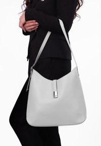 Šedá kožená kabelka přes rameno s hladkým povrchem, minimalistickým designem a kovovým zapínáním, nesená na rameni.