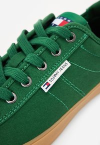 Green canvas sneaker with round metal eyelets, flat laces, and a gum sole. Features a white label with "TOMMY JEANS" and a logo accent.