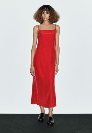 Red satin slip dress with thin straps, floor-length design, smooth texture, worn with black clogs on a dark surface against a light background.