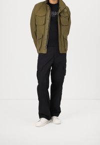 Let olivengrøn jakke i letvægtsmateriale med flere lommer, sort t-shirt, sorte cargobukser og hvide sneakers. Casual og funktionelt design.