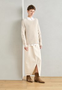 Suéter de punto beige con mangas largas, superpuesto a una camisa blanca de botones, combinado con una falda color crema y botas de tobillo de ante marrón.
