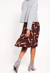 Pull court gris sur une jupe à motifs avec des rayures en chevron rouge, orange et marine ; assorti à des bottines grises.