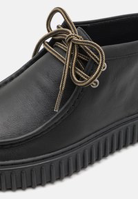 Chaussure en cuir noir avec un bout rond et une semelle en caoutchouc texturé. Présente des lacets en jute beige et des œillets en métal sur le dessus.