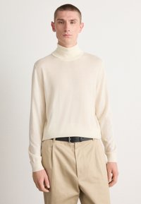 Pull à col roulé en crème en tricot léger avec des manches longues, associé à un pantalon kaki et une ceinture noire, présentant une texture lisse.