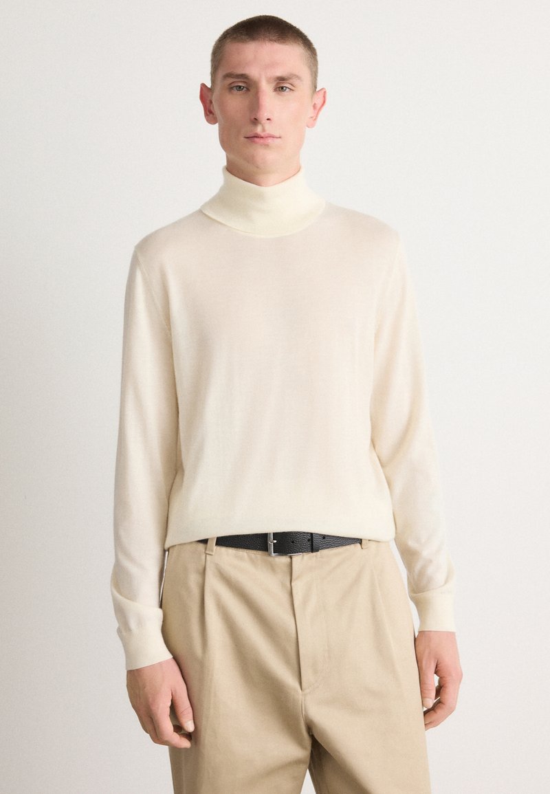 Pull à col roulé en crème en tricot léger avec des manches longues, associé à un pantalon kaki et une ceinture noire, présentant une texture lisse.