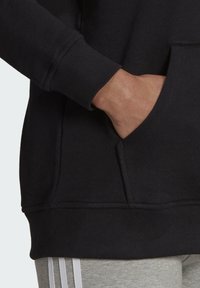 Felpa nera con polsini a costine e tasca frontale. La texture sembra morbida con un orlo spesso, mostrata insieme a pantaloni grigi con strisce laterali.