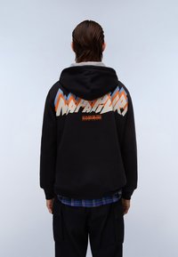 Felpa nera con cappuccio presenta un design colorato di montagne e il nome del marchio "Napapijri" sulla schiena. Indossata sopra una camicia a quadri.
